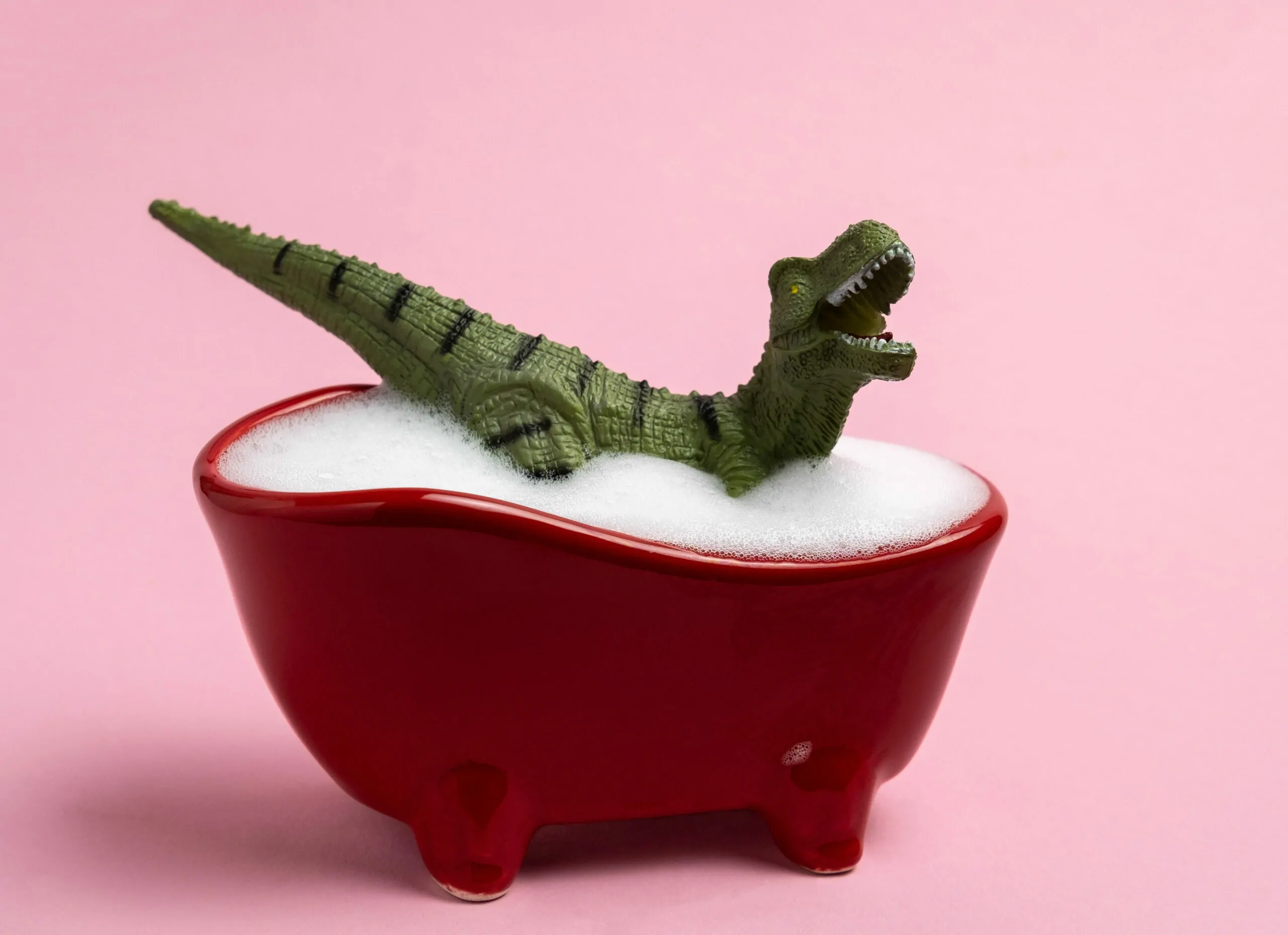 Cute dinosaur toy washing in red bath with foam on pink background.